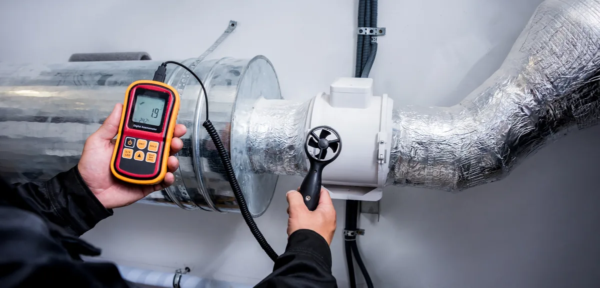 Hands holding an airflow meter beside insulated ventilation ducts